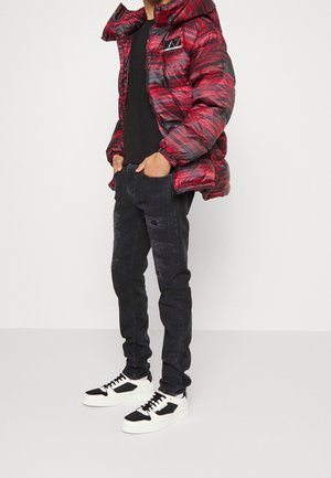 Red and gray patterned puffer jacket, black t-shirt, distressed black jeans with textured patches, and white sneakers with black accents.