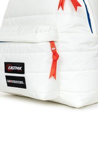 Bílý polstrovaný batoh s červenými akcenty, včetně zipů a mašlí. Zobrazuje značkové nášivky "EASTPAK" a "GHOSTBUSTERS". Texturovaný povrch.