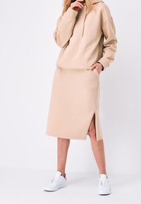Sweat à capuche beige avec cordon de serrage et manches longues, associé à une jupe beige longueur genou avec une fente latérale, porté avec des baskets blanches.