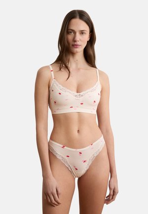 Frau trägt einen hellrosa BH mit Spitzenbesatz und passende Unterhose mit rotem und rosa Lippenstiftmuster vor weißem Hintergrund.