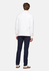 Sudadera blanca de manga larga, con puños y dobladillo acanalados, combinada con vaqueros azul oscuro. Modelo visto desde atrás, con zapatillas casuales.