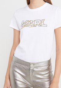 Valkoinen puuvillainen T-paita, jossa on mustalla ääriviivalla varustettu "KARL"-logo kultaisilla koristeilla. Malli yhdistää sen metallinhohtoisiin hopeisiin housuihin.