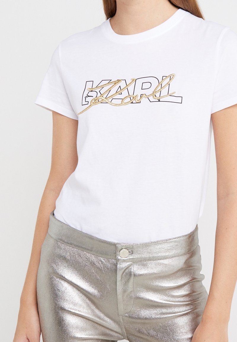 Valkoinen puuvillainen T-paita, jossa on mustalla ääriviivalla varustettu "KARL"-logo kultaisilla koristeilla. Malli yhdistää sen metallinhohtoisiin hopeisiin housuihin.