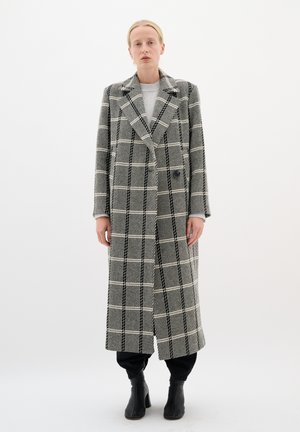 Lungo cappotto con motivo a quadri in bianco e nero, con ampi revers e due bottoni, abbinato a pantaloni neri e stivaletti alla caviglia.