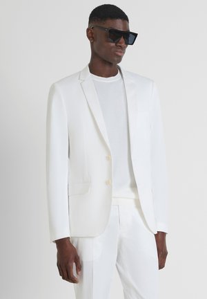 Wit kostuum met een op maat gemaakte jas en bijpassende pantalon. Glad, glanzend materiaal. Gecombineerd met een wit overhemd en grote zwarte zonnebril.