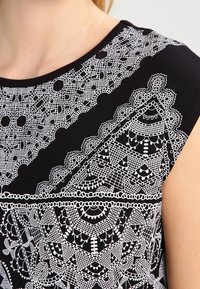 Schwarzes Oberteil mit aufwendigen, weißen Spitzenmustern, geometrischen Designs und einer glatten Textur, mit rundem Ausschnitt und kurzen Ärmeln.