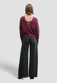 Suéter de punto burdeos con detalle de cordones en la espalda, combinado con vaqueros negros de pierna ancha. El conjunto presenta un acabado texturizado y espalda abierta.