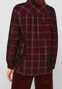 Camicia flanella a quadri rossi e neri con maniche lunghe, orlo arrotondato e cucitura sulla spalla posteriore. Texture morbida con accenti bianchi tenui.