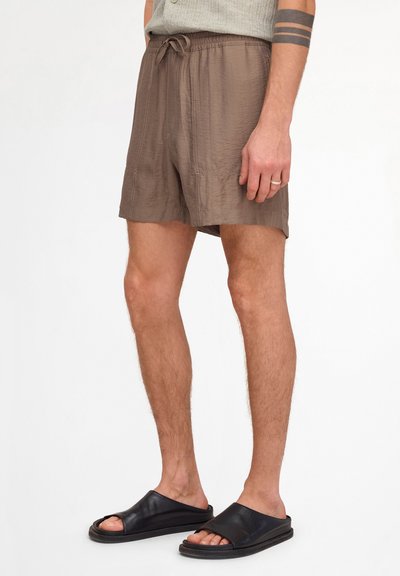 Homme portant un short marron avec cordon, un haut gris sans manches, des sandales noires à enfiler et un tatouage noir sur le bras avec deux bandes épaisses.