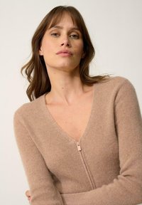 Cárdigan de punto color beige con un profundo cuello en V, mangas largas y cremallera central. Textura suave con un sutil detalle de canalé.