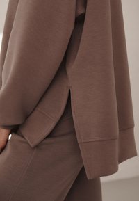 Felpa marrone con spacco laterale, tessuto morbido, vestibilità comoda e polsini a costine. Dettaglio delle cuciture visibili lungo l'orlo e le maniche.