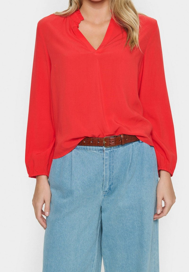 Femme portant un chemisier rouge ample à manches longues, rentré dans un jean taille haute bleu clair avec une ceinture marron.