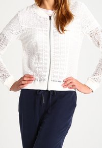 Bílá pletená bomber bunda na zip s otevřenou tkaninou a pružnými manžetami. V kombinaci s tmavě modrými kalhotami.