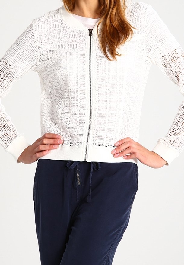 Bílá pletená bomber bunda na zip s otevřenou tkaninou a pružnými manžetami. V kombinaci s tmavě modrými kalhotami.