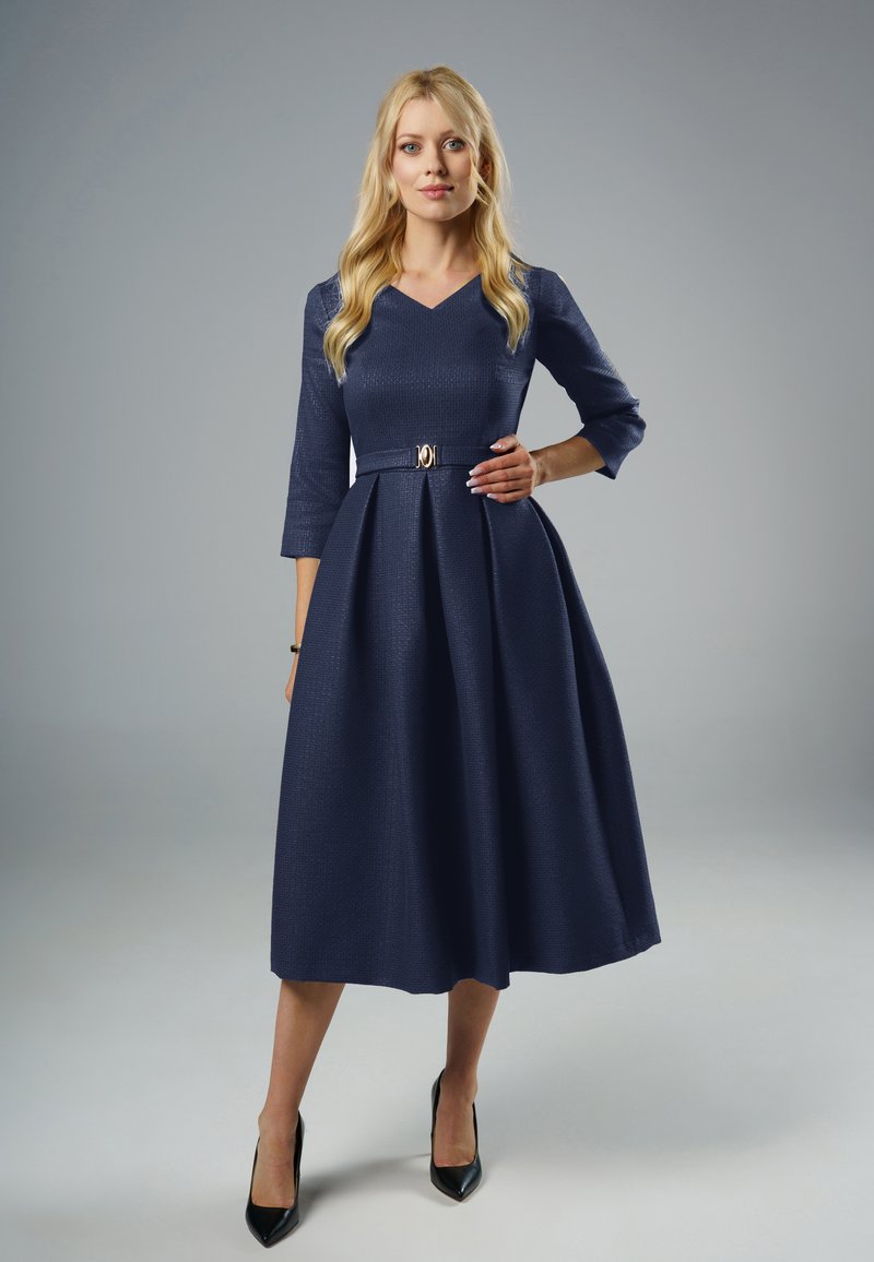 Granatowa sukienka z rękawem do trzech czwartej, w dekolcie w kształcie litery V, z zaznaczoną talią i plisowanym dołem; ma złotą klamrę paska oraz teksturowany materiał.