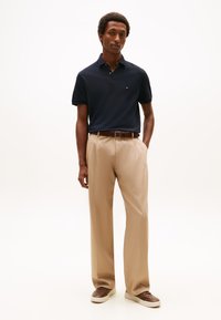 Homem de pé vestindo uma camisa polo azul-marinho, calças bege com um cinto castanho e ténis castanhos contra um fundo branco.