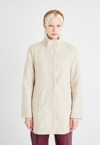 Beige ullblandad kappa med hög krage, rak skärning, sidofickor och subtila sömdetaljer. Bärs över en ljusrosa polotröja.