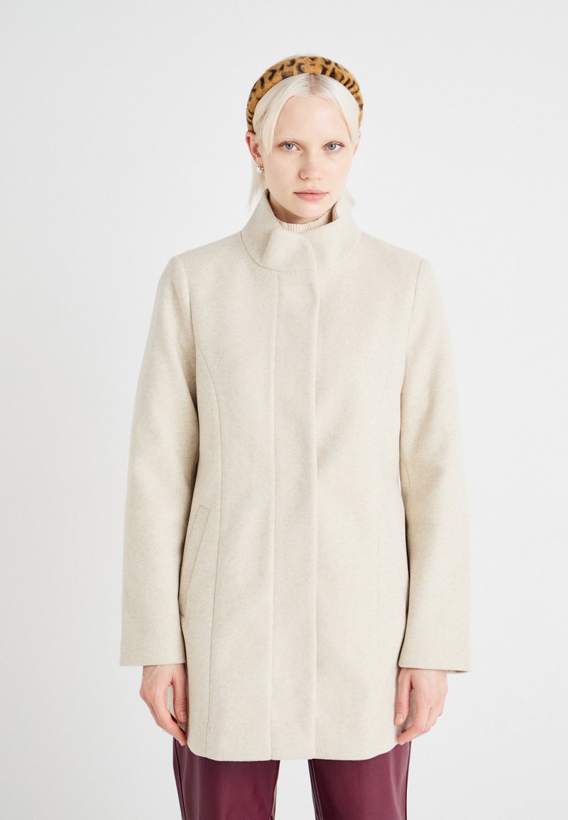 Beige ullblandad kappa med hög krage, rak skärning, sidofickor och subtila sömdetaljer. Bärs över en ljusrosa polotröja.