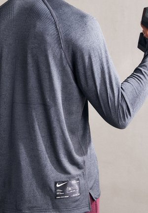 Persoon draagt een grijs Nike langarm sportshirt met een label en houdt een zwarte dumbbell in hun rechterhand tegen een effen achtergrond.
