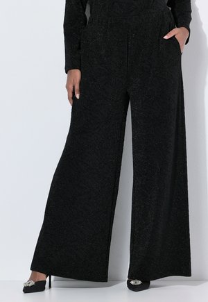 Persoon draagt zwarte wijde broek met subtiele patronen en glitters, één hand in de zak, gecombineerd met zwarte puntige hoge hakken met decoratieve broches.
