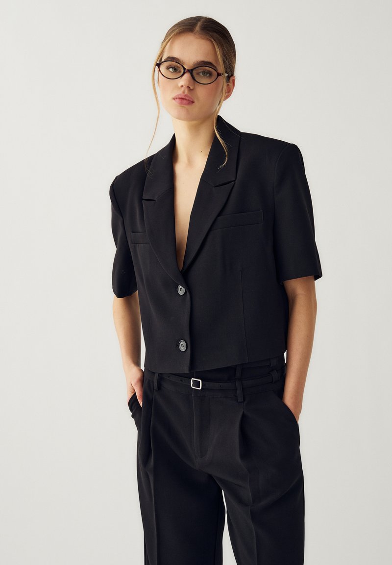 Joven con gafas que lleva un blazer negro de manga corta y pantalones negros a juego de cintura alta, de pie con las manos en los bolsillos.