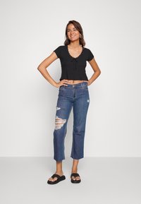 Top preto curto com decote em zig-zag e laço frontal, combinado com jeans azuis rasgados e sandálias pretas de slide.