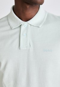 Svetlozelená polo tričko vyrobené z textúrovanej tkaniny, s klasickým golierom, tromi gombíkmi a decentným vyšitým logom BOSS v svetlomodrej farbe.