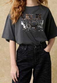 Gray short-sleeve T-shirt with sequin lettering “LET IT SNOW” in shades of silver and bronze; paired with black patterned denim jeans.