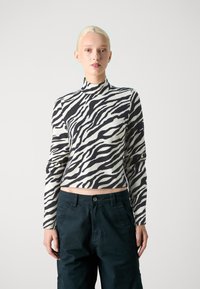 Cropped top s crno-bijelim prugama poput zebre, s visokim ovratnikom i dugim rukavima, u kombinaciji s hlačama tamnozelene boje sa širokim pojasom, prikazajući glatku teksturu tkanine.
