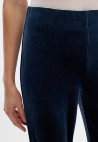 Tumši zila samta legingi ar gludu tekstūru, augstu jostu un šauru piegriezumu. Redzama roka, kas tur audumu, akcentējot tā mīkstumu.