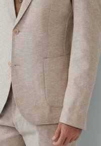 Beige linnen colbert met drie knopen, één zak aan de linkerkant, gestructureerde stof, getailleerde pasvorm, zichtbare stikseldetails.