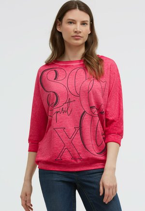 Frau trägt ein knallpinkes Langarmshirt mit großen schwarzen Buchstaben und den Worten „spirit“ und lässige blaue Jeans.