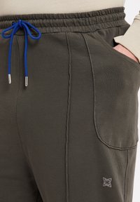 Olive green joggers with a textured fabric, elastic waistband, blue drawstring, side pockets, and a subtle logo accent on the hip.