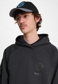 Svart huvtröja med grå detaljer, med en logotyp på bröstet. Kapseln är svart med en grå skärm och blå logotydelse.