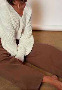 Vit stickad cardigan med V-ringning, med strukturerade mönster och tre stora knappar, i kombination med bruna vida byxor på ett betonggolv.