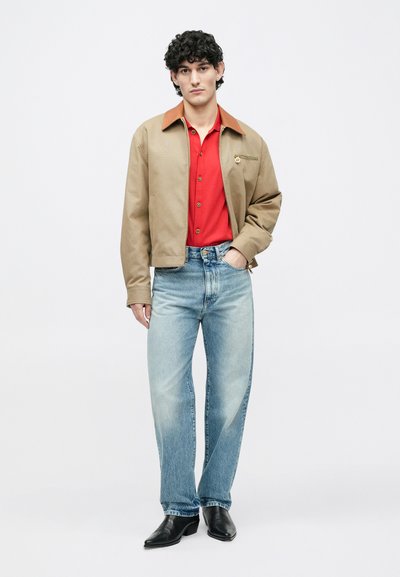 Beige Bomberjacke mit braunem Kragen, rotem Poloshirt, hellblauen Jeans und schwarzen Schuhen. Die Jacke verfügt über eine Fronttasche und einen Reißverschlussdetail.