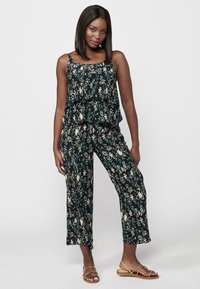 Combinaison noire à motif floral avec un haut à volants superposés, pantalon large et bretelles spaghetti fines. Accessoires complétant le look : sandales dorées.