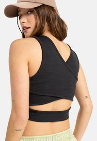 Sort cropped top med tekstureret stof, med et krydset rygdesign; parret med lyse bukser og en beige kasket.
