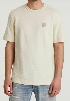 T-shirt crème à manches courtes avec des rayures texturées, col rond et petite étiquette avec logo sur la poitrine. Porté avec un jeans bleu déchiré.