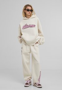 Felpa oversize color crema con ricami rosa, abbinata a pantaloni della tuta coordinati con dettagli simili. Indossata con sneakers marroni e rosa.