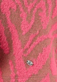 Primer plano de una tela rosa con estampado, textura difusa y una pequeña placa ovalada plateada grabada con un corazón y las palabras "Key Largo".