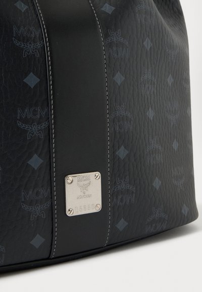 Gros plan d’un sac en cuir noir MCM avec des logos à motifs, une bande verticale en cuir et une plaque de marque argentée avec des vis et un numéro de série.