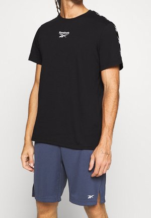 Mann trägt ein schwarzes Reebok-T-Shirt und blaue Reebok-Shorts und steht vor einem einfarbigen weißen Hintergrund.