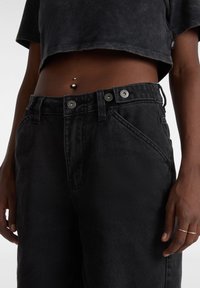Pantaloni neri in denim a vita alta con due bottoni argentati, tasche angolate e una consistenza morbida. Top corto visibile sopra la vita.