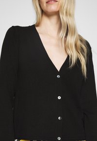 Czarny sweter na drutach z dekoltem w serek, długimi rękawami i rzędem srebrnych guzików z przodu. Gładka faktura i dopasowany krój.