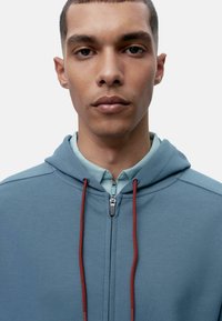 Svijetloplava zip-up dukserica napravljena od mekog materijala, s crvenim špagicama i bijelom košuljom s kragnom ispod.