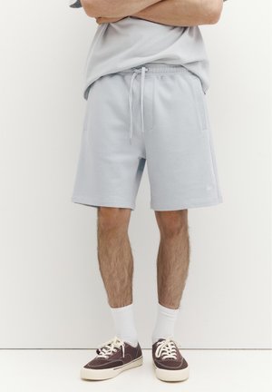 Bărbat purtând pantaloni scurți gri deschis cu șnur, șosete albe și adidași maro închis, stând cu brațele încrucișate pe un fundal alb simplu.