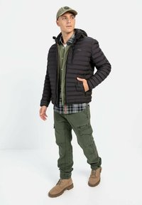 Sort pufferjakke med vandret søm, grøn skjorte med knapper, grøn cargo-bukser, lyse brune støvler og en lys kasket. Synlige sidelommer.