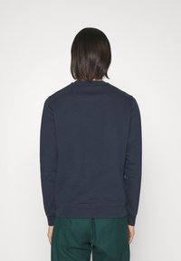 Person med axellångt mörkt hår som bär en marinblå sweatshirt och gröna byxor, stående mot en enkel vit bakgrund.