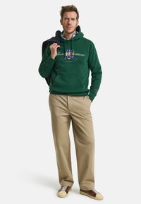 Green hoodie with a front pocket and graphic design. Beige pants and brown sneakers with cream accents. Model holding a navy blazer.